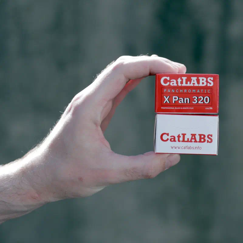 A hand holding a white CatLABS X Pan 320 carton, lettered in red and marked PANCHROMATIC, against a grey wall