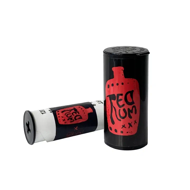 A 120 roll of CineStill REDRUM standing on end beside a second roll on its side, black wrapper with the name hand-lettered in red inside a bottle