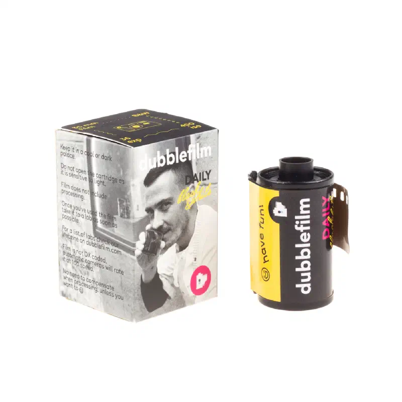 A dubblefilm DAILY carton standing beside its 35 mm cassette, the carton printed with a black-and-white portrait of a man at a telephone and a magenta dot, the cassette in dubblefilm's yellow and black