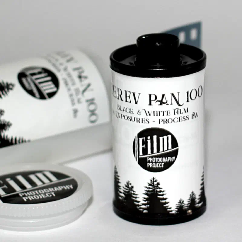 A 35 mm cassette of DEREV PAN 100, white with a black band of fir trees around the base and the Film Photography Project roundel, a second cassette lying behind it