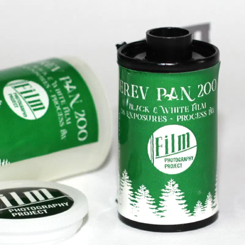 A 35 mm cassette of DEREV PAN 200, green with a white band of fir trees around the base and the Film Photography Project roundel, a second cassette lying behind it