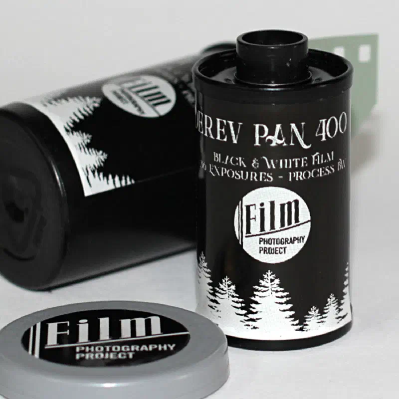 A 35 mm cassette of DEREV PAN 400, black with a white band of fir trees around the base and the Film Photography Project roundel, a second cassette lying behind it