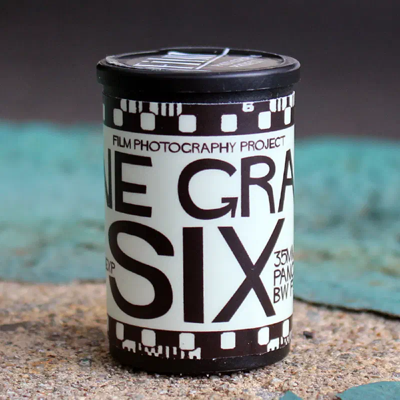 A 35 mm canister standing on stone, black and white with a sprocket-hole border, lettered FINE GRAIN SIX under the Film Photography Project roundel