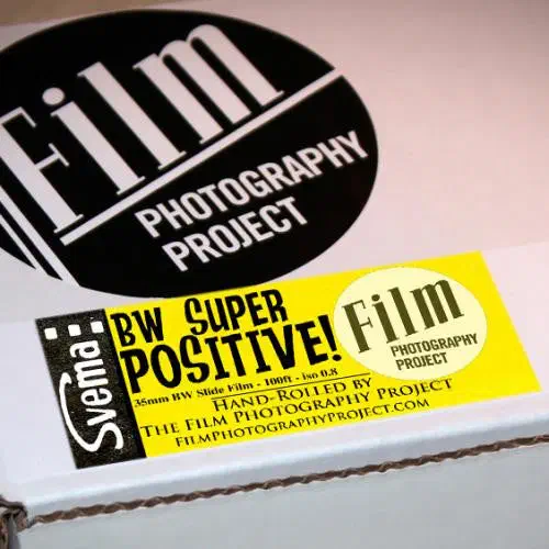 A white Film Photography Project box seen from a front corner, its yellow label reading Svema BW SUPER POSITIVE!, 35mm BW Slide Film, hand-rolled by The Film Photography Project