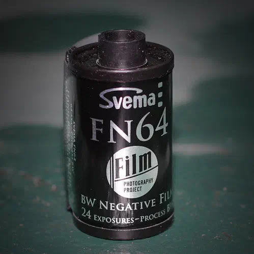 A 35 mm cassette of Svema FN 64 standing on a green surface, silver label marked BW NEGATIVE FILM, 24 EXPOSURES