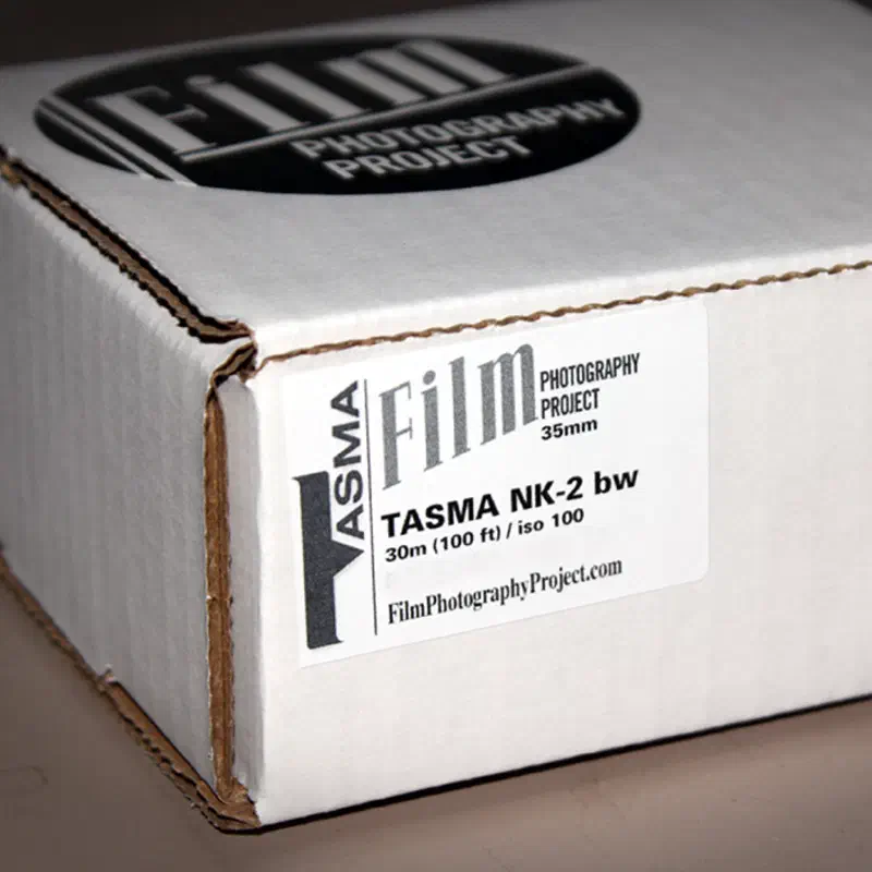 A white Film Photography Project box seen from a front corner, a small label at the front reading TASMA NK-2 bw, 30m (100 ft) / iso 100