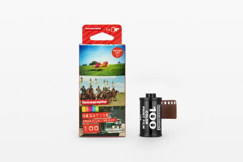 A Lomography Color Negative 100 three-roll carton printed with sample frames, beside a black 35 mm cassette and leader, marked 35MM FILM / 36 EXP