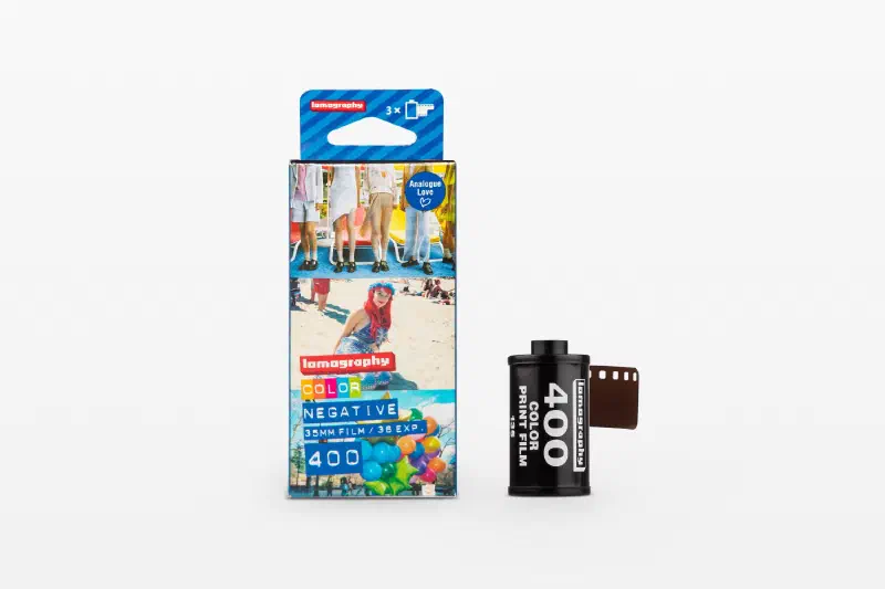 A Lomography Color Negative 400 three-roll carton printed with sample frames, beside a black 35 mm cassette and leader, marked 35MM FILM / 36 EXP