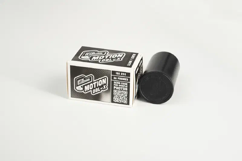 A small black-and-white Optik's MOTION DBL-X carton standing beside a black film tin on a plain ground