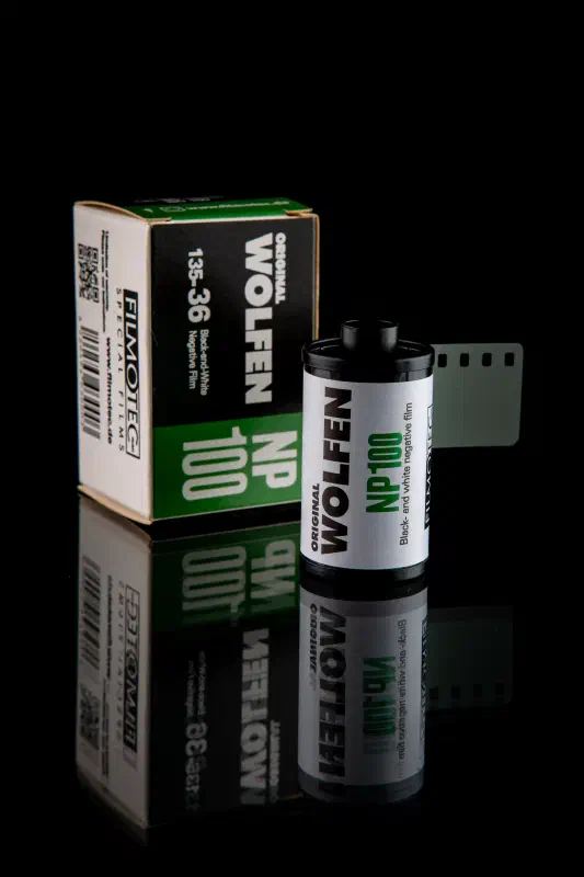 A WOLFEN NP100 carton standing on black beside its 35 mm cassette, both white with a green band, the film leader out