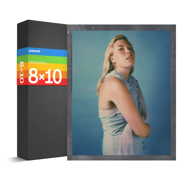 A black Polaroid 8×10 box seen from a front corner, its rainbow stripe across the face, a developed portrait of a woman in a blue dress leaning against it