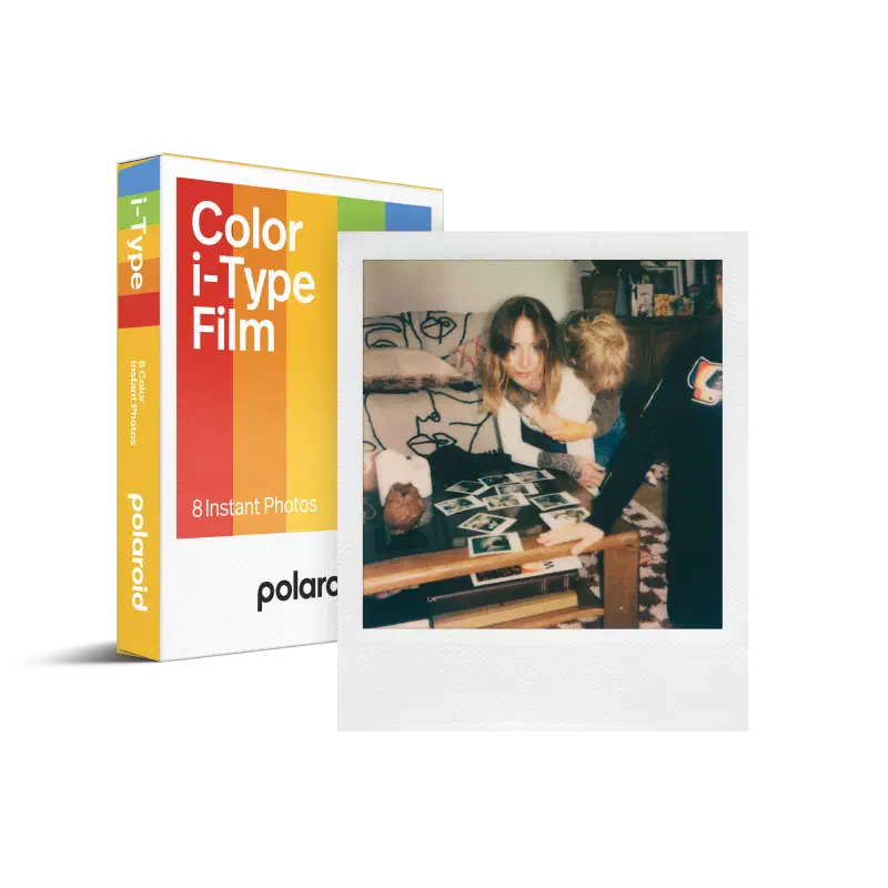 A white Polaroid Color i-Type Film box seen from a front corner, its rainbow stripe across the face and 8 Instant Photos at the foot, a developed picture of two people at a table leaning against it