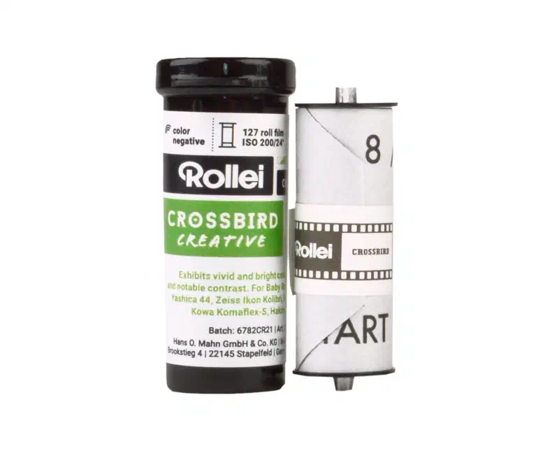 A 127 roll of Rollei CROSSBIRD CREATIVE, its white wrapper lettered in green and black and marked color negative, 127 roll film, ISO 200/24, standing beside a second roll unwound to show the numbered backing paper